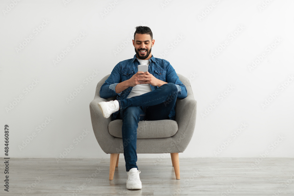 Cheerful young Arab man using smartphone, having online chat, sitting in armchair, browsing social media