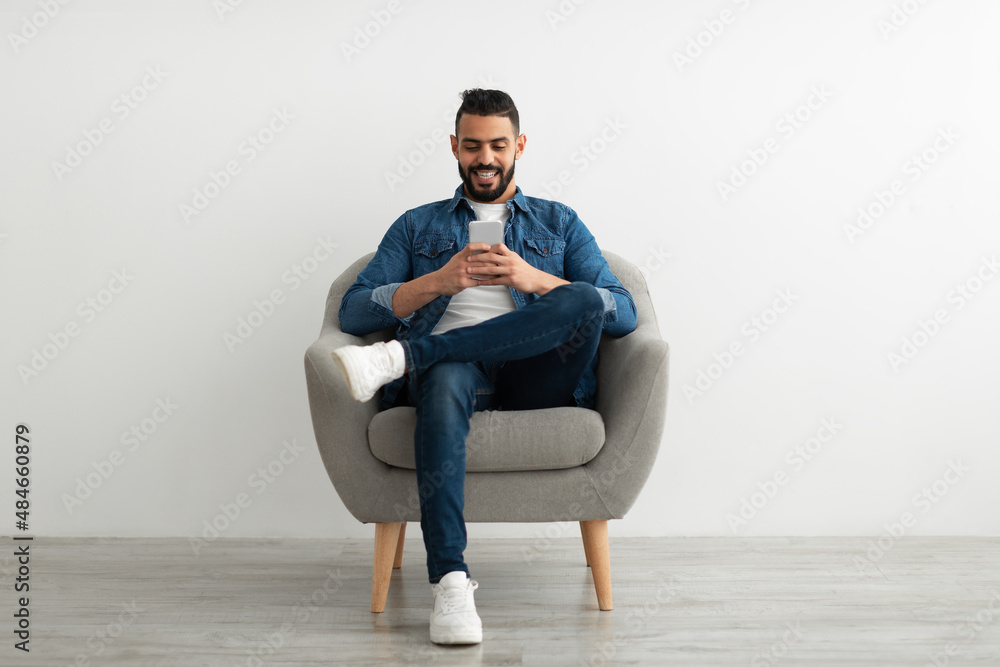 Cheerful young Arab man using smartphone, having online chat, sitting in armchair, browsing social media Cheerful young Arab man using smartphone, having online chat, sitting in armchair, browsing social media