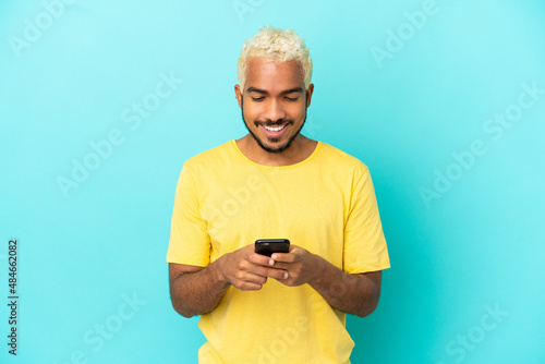 Young Colombian handsome man isolated on blue background sending a message with the mobile © luismolinero