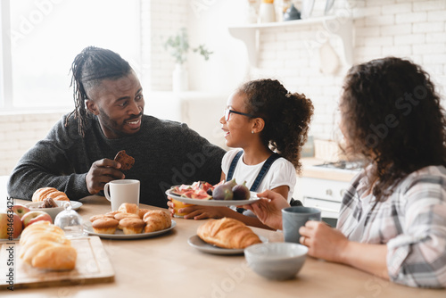 Healthy eating. African-american family  parents and daughter having morning breakfast at home kitchen  talking and spending time together. Family bonding