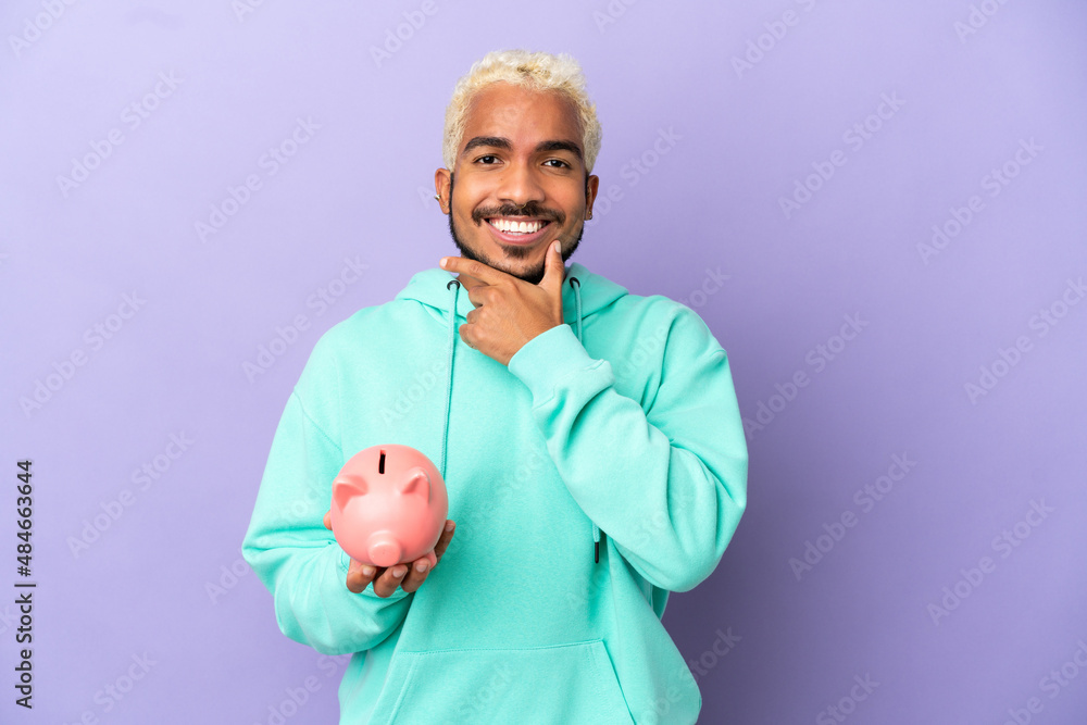 Young Colombian man holding a piggybank isolated on purple background happy and smiling
