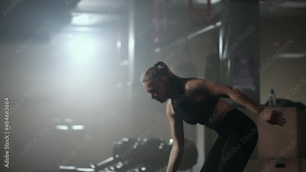 In slow motion, a woman lifts a dumbbell over her head while working out in a dark gym. Athletic strong woman performs a difficult workout with lifting dumbbells In slow motion, a woman lifts a dumbbell over her head while working out in a dark gym. Athletic strong woman performs a difficult workout with lifting dumbbells