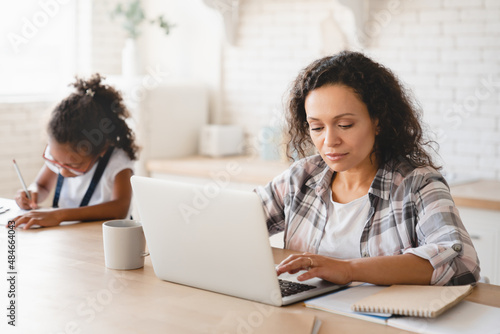 Maternity leave. Social distancing and stay at home. African mother mom freelancer working remotely on laptop while daughter doing homework, painting at home kitchen. E-learning