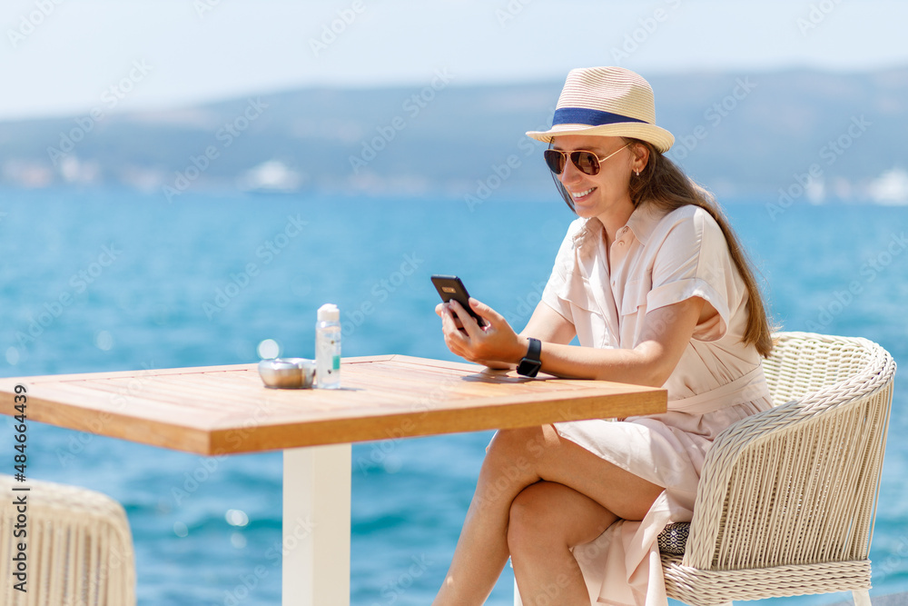 Young lady in dress sitting at the table in cafe near the sea. Young travel woman enjoying sunny day at restaurant on her vacation