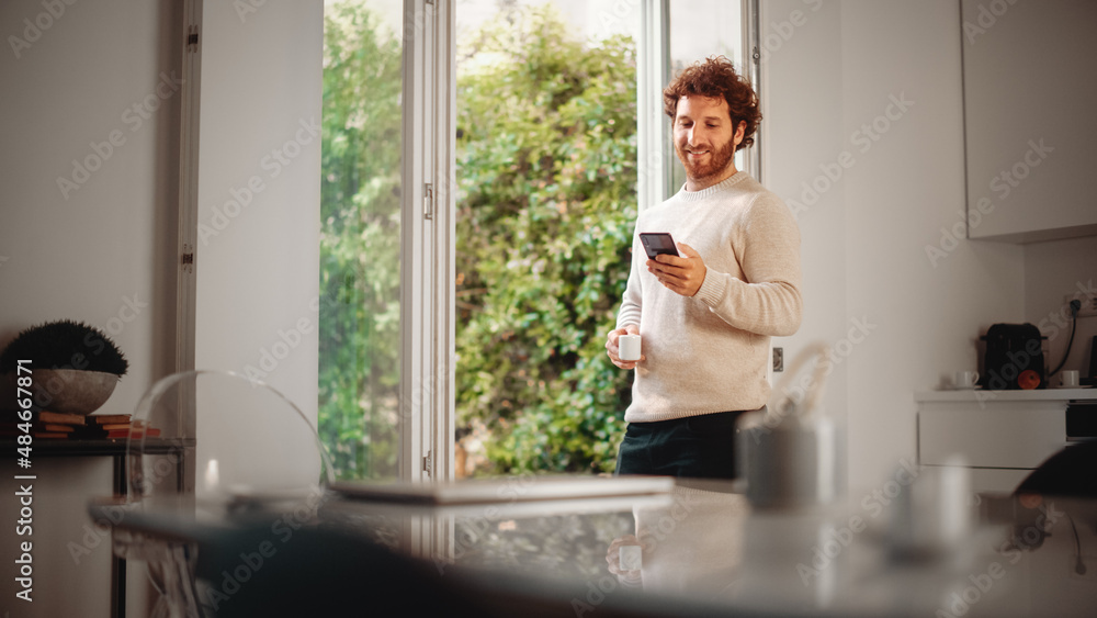 Handsome Adult Man with Ginger Curly Hair Using Smartphone, Standing in Living Room at Home. Male Enjoys a Cup of Espresso, Checking Social Media, Online Shopping, Watching Videos or Writing Messages.