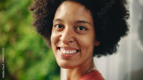 Close Up Portrait of a Happy Young Latina with Brown Eyes, Afro Hair and Nose Piercing Posing for Camera. Beautiful Diverse Multiethnic Black Hispanic Female Smiling on Green Nature Background. © Gorodenkoff