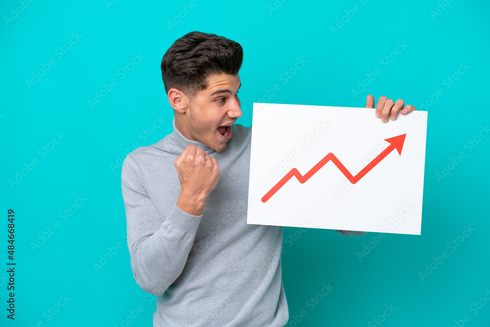 Young handsome caucasian man isolated on blue bakcground holding a sign with a growing statistics arrow symbol and celebrating a victory Young handsome caucasian man isolated on blue bakcground holding a sign with a growing statistics arrow symbol and celebrating a victory