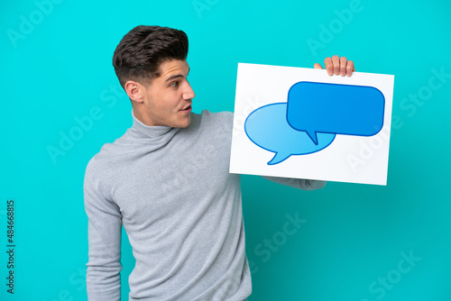 Young handsome caucasian man isolated on blue bakcground holding a placard with speech bubble icon