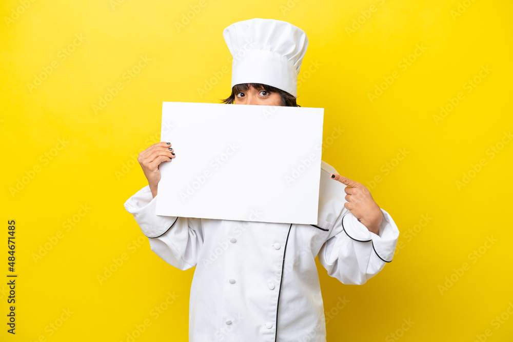 Young chef latin woman isolated on yellow background holding an empty placard and hiding behind it Young chef latin woman isolated on yellow background holding an empty placard and hiding behind it