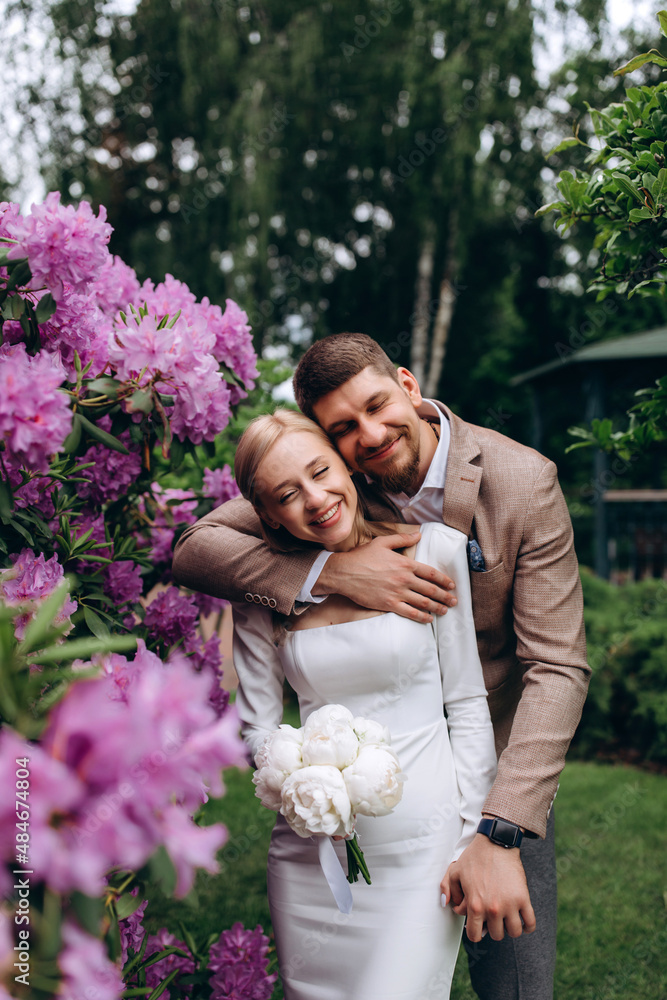 Walk of the bride and groom in the summer park. The bride hugs the groom. Photo on a background of pink flowers. Newlyweds in seclusion. Walk of the bride and groom in the summer park. The bride hugs the groom. Photo on a background of pink flowers. Newlyweds in seclusion.