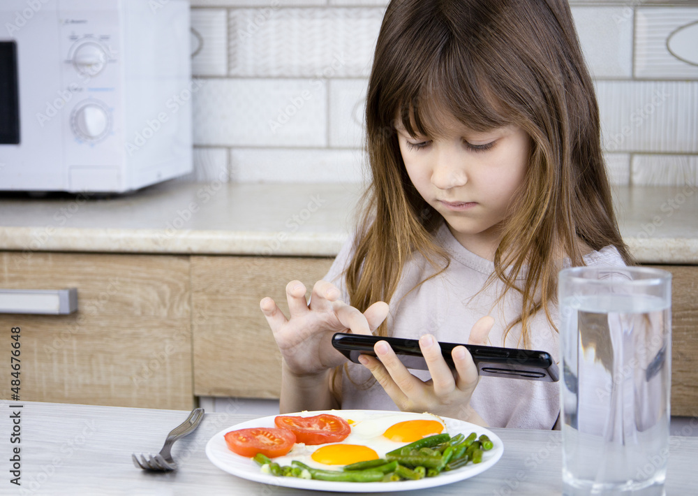 The child does not want to eat breakfast.The girl in the kitchen refuses to eat and plays on the phone.Internet addiction.A child and a gadget. Social problems.Fried eggs, tomatoes and beans on a plat