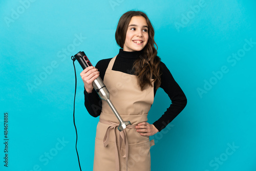 Little girl using hand blender isolated on blue background posing with arms at hip and smiling