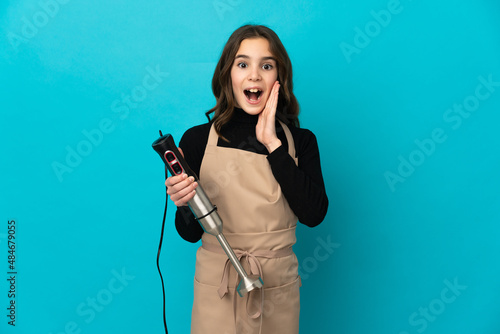 Little girl using hand blender isolated on blue background with surprise and shocked facial expression