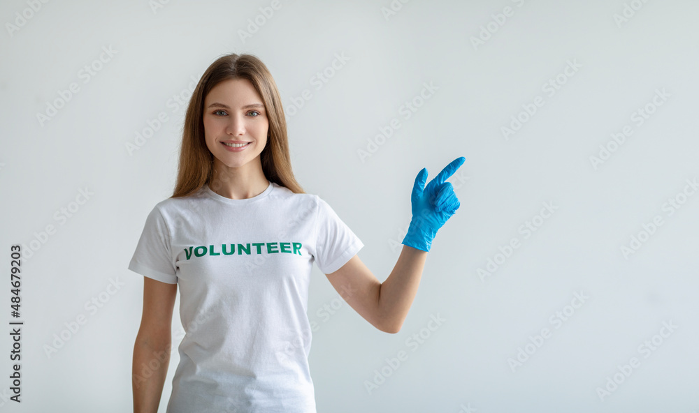 Look here. Happy female volunteer in gloves pointing aside at free space, standing over light wall and smiling at camera Look here. Happy female volunteer in gloves pointing aside at free space, standing over light wall and smiling at camera