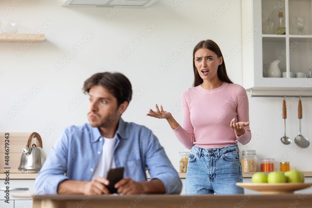 Sad angry upset offended young wife yelling at husband with smartphone on kitchen interior