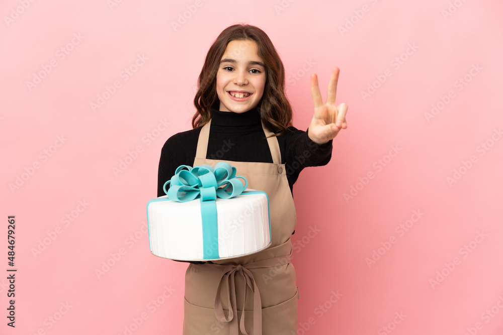 Little Pastry chef holding a big cake isolated on pink background smiling and showing victory sign Little Pastry chef holding a big cake isolated on pink background smiling and showing victory sign