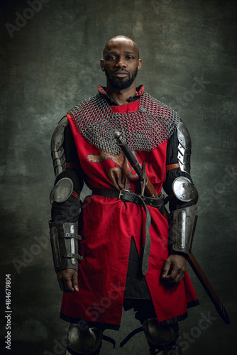 Vintage style portrait of brutal dark skinned man, medieval warrior or knight with wounded face wearing armour isolated over dark background. Comparison of eras, history © master1305