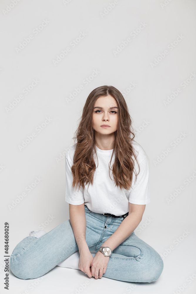 young pretty girl with clean skin and a hairstyle in a white T-shirt with  fashionable blue jeans sits on a white background in the studio Stock Photo  | Adobe Stock
