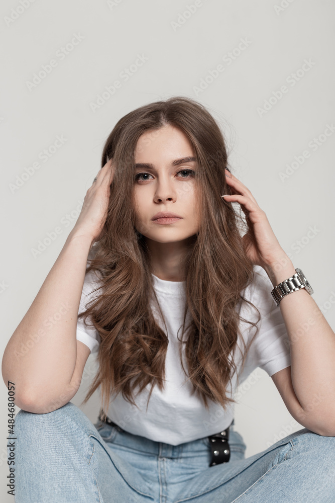 Fashionable portrait of a beautiful girl with make-up and hairstyle in a  white T-shirt and blue jeans sits in the studio Stock Photo | Adobe Stock