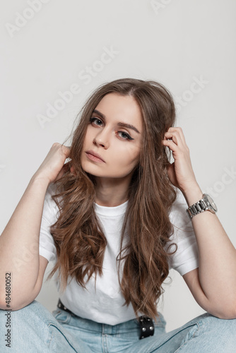 Pretty fashion seriously woman model in white t-shirt and blue jeans sits in studio on a white background. Beauty girl with hairstyle in stylish summer outfit