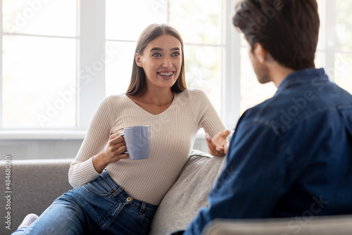 Happy emotional millennial caucasian woman with cup of drink sit on couch, gesturing, rest and communicating