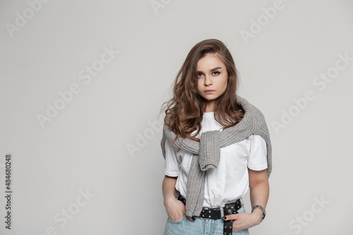 Cool stylish young woman model in a fashionable casual outwear with a knitted sweater, white T-shirt and blue jeans stands in the studio on a white background. Female outfit © alones