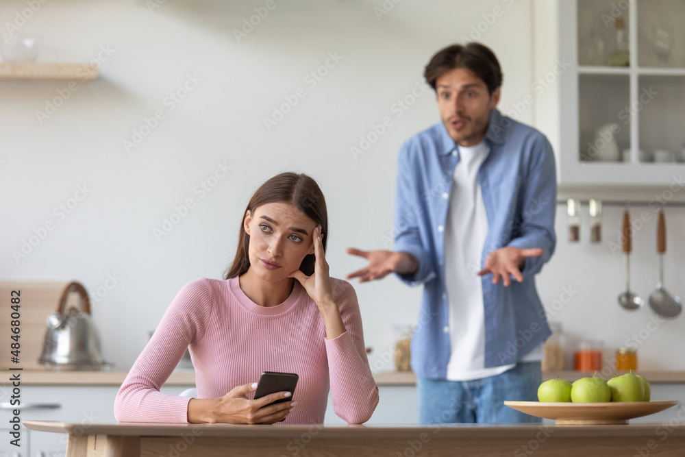 Depressed sad young caucasian lady with phone ignoring frustrated scolding man in kitchen interior Depressed sad young caucasian lady with phone ignoring frustrated scolding man in kitchen interior