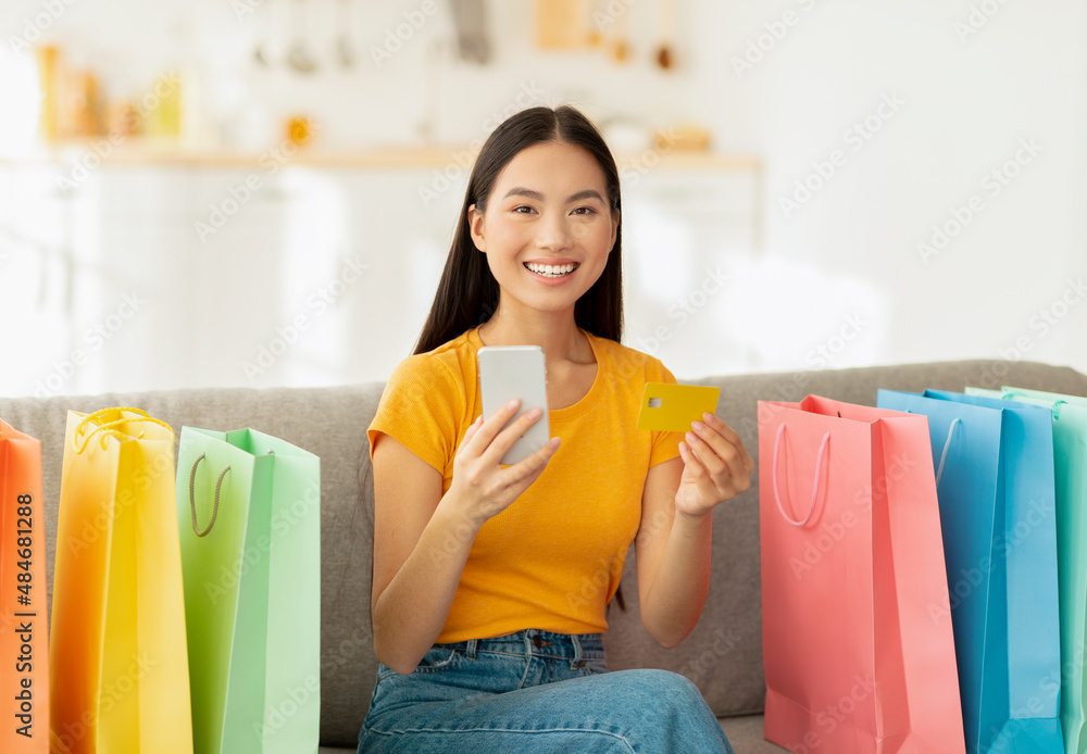 Positive asian lady with credit card and smartphone sitting on sofa surrounded by colorful shopping bags Positive asian lady with credit card and smartphone sitting on sofa surrounded by colorful shopping bags