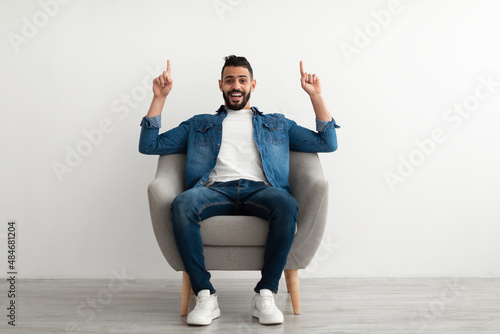 Full length of joyful young Arab guy pointing upwards with both hands, sitting in armchair against white studio wall