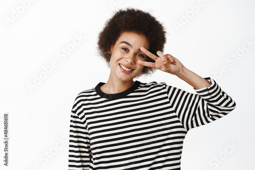 Positive Black girl with afro hair, smiling and looking healthy, showing peace v-sign salute, standing over white background