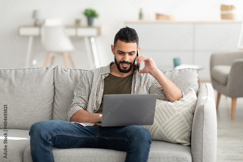 Handsome millennial Arab man working on laptop and calling on smartphone, having remote job, indoors Handsome millennial Arab man working on laptop and calling on smartphone, having remote job, indoors
