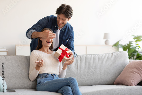 Smiling young european husband closes eyes to his wife and gives gift box in living room interior photo