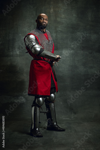 Portrait of cheerful dark skinned man, medieval warrior or knight with wounded face wearing armour making faces isolated over dark background. Comparison of eras, history