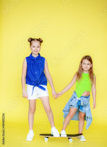 Funny little blonde girls 6-10 years old in everyday bright clothes pose isolated on the yellow background of the studio. The concept of a child's lifestyle. Layout of the copy space. Hold the skatebo