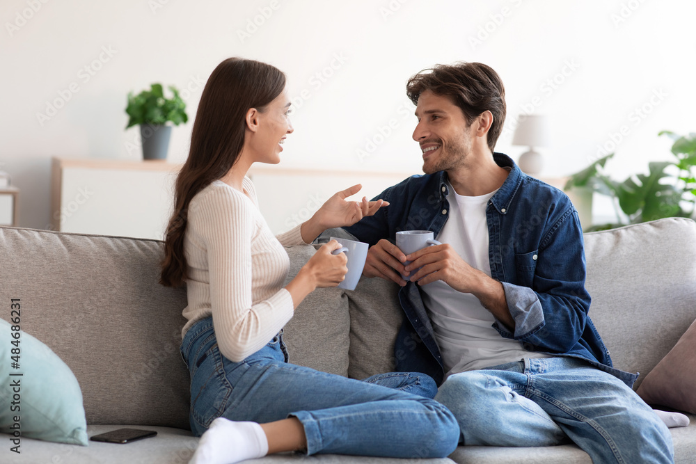 Smiling millennial caucasian male and female sit on sofa with cups of hot drink and talk in living room Smiling millennial caucasian male and female sit on sofa with cups of hot drink and talk in living room