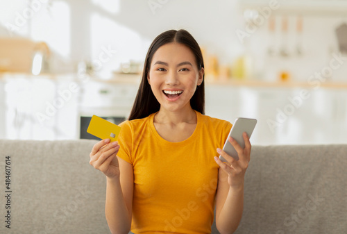 Cybershopping, seasonal sales concept. Overjoyed asian lady with cellphone and credit card buying goods in online store photo