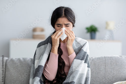 Unhappy sad young indian female in plaid suffering from fever and flu on sofa, blowing nose in napkin photo