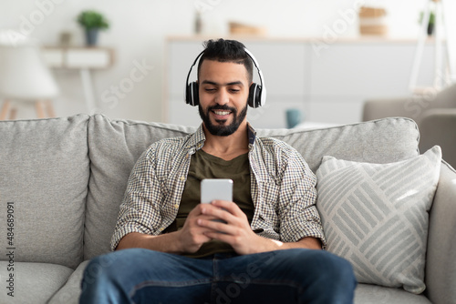 Cheerful young Arab guy watching movie while resting on couch at home, using wireless headset and mobile phone