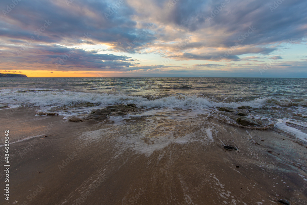 Golden hour sunset at Whitby beach. Warm sky and gentle, smooth waves along a sandy coastline with large rocks.