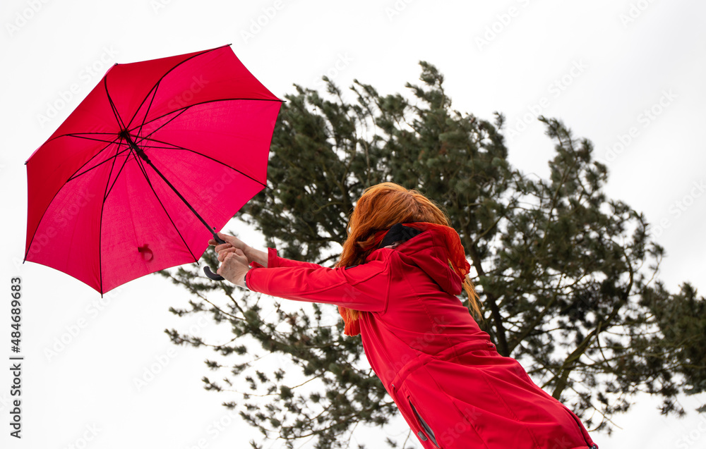 Eine Frau versucht bei starkem Wind ihren roten Regenschirm festzuhalten.  Im Hintergrund steht ein Baum. Stock Photo | Adobe Stock