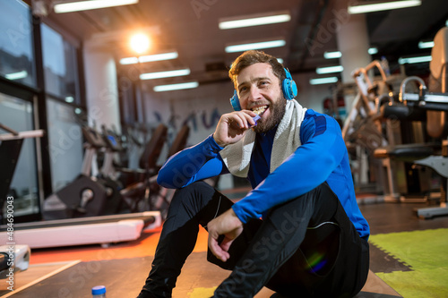 A smiling guy sits after a workout in the gym, listens to music on headphones and eats a protein bar