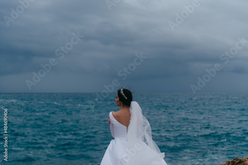 Back view of brunette bride in white wedding dress and bridal veil on a cloudy day. Romantic beautiful bride posing near the sea with waves