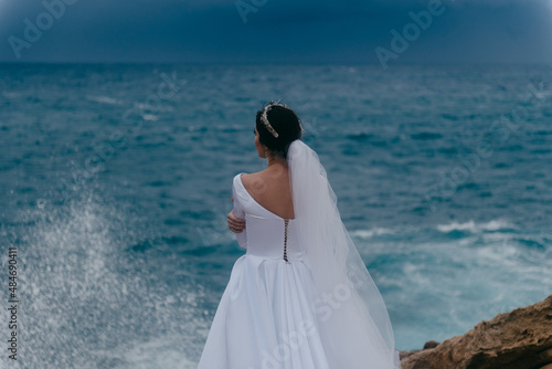Back view of brunette bride in white wedding dress and bridal veil on a cloudy day. Romantic beautiful bride posing near the sea with waves