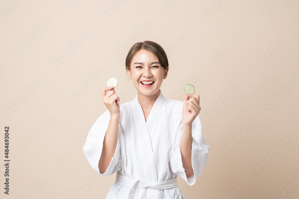 Portrait Beautiful young Asian women happy smile in bathrobe or spa suit with holding cucumber slice for facial face mask on brown isolated studio background. Portrait Beautiful young Asian women happy smile in bathrobe or spa suit with holding cucumber slice for facial face mask on brown isolated studio background.