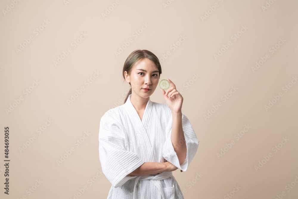 Portrait Beautiful young Asian women in bathrobe or spa suit with holding cucumber slice for facial face mask on brown isolated studio background. Portrait Beautiful young Asian women in bathrobe or spa suit with holding cucumber slice for facial face mask on brown isolated studio background.