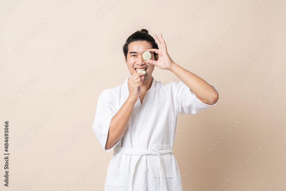 Portrait Asian man happy smile in bathrobe or spa suit with holding cucumber slice for facial face mask on brown isolated studio background.