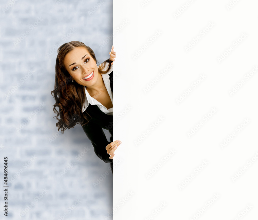 Happy smiling business woman, standing behind, peeping from blank banner or mock up signboard, showing copy space, on white bricks loft wall background Happy smiling business woman, standing behind, peeping from blank banner or mock up signboard, showing copy space, on white bricks loft wall background