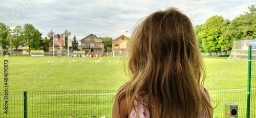 Sremska Mitrovica, Serbia, August 1, 2020. A girl sits with her back to the camera and watches a football match of childrens teams. Beautiful long blonde hair. Soccer fans in a match photo