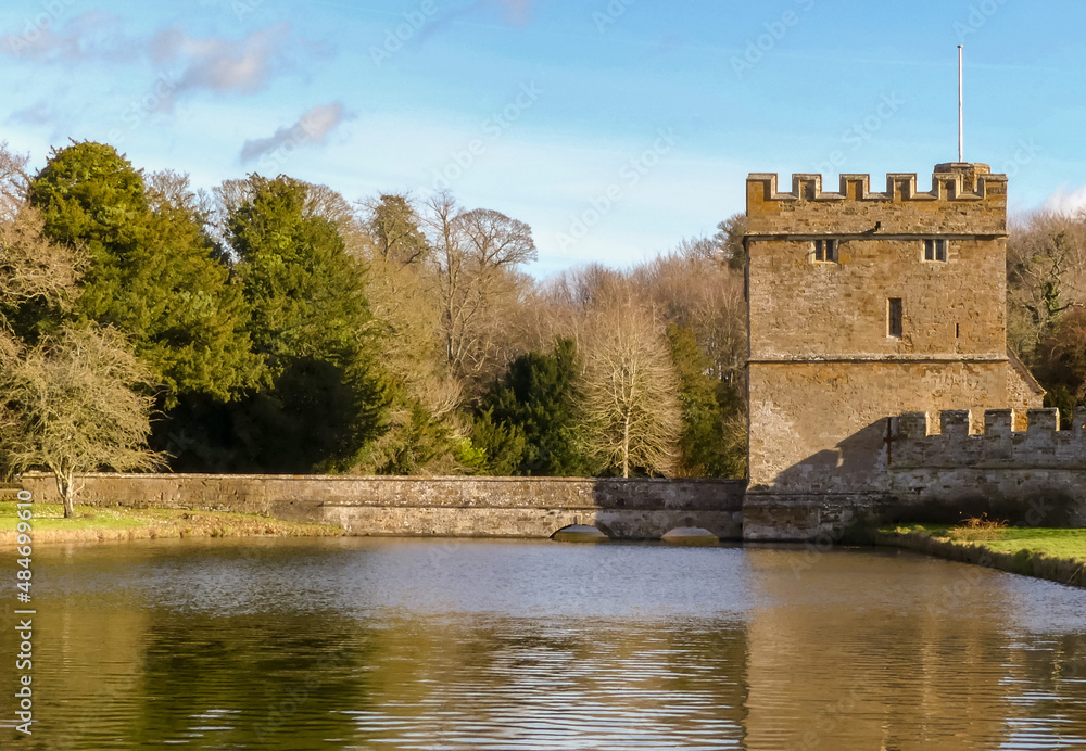 Public view of a medieval castellated gatehouse with bridge access over a moat. Water gently rippling. Woodland background under a blue and clouded sky. Landscape image with space for text. England. Public view of a medieval castellated gatehouse with bridge access over a moat. Water gently rippling. Woodland background under a blue and clouded sky. Landscape image with space for text. England.