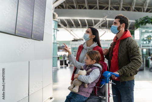 Side view of the parents wearing protective masks standing in front of the timetable board and waiting for the flight at the airport while their daughter pointing with finger. 
